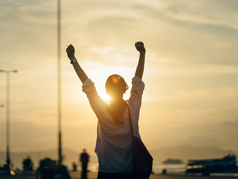 A woman with a bag on shoulder, facing towards sun and raised her hands